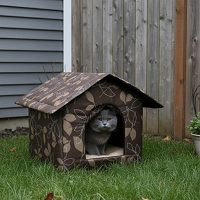 Niche pour chat extérieur en tissu motif feuilles avec chat gris russe regardant depuis l'entrée, placée sur gazon près d'une clôture en bois.